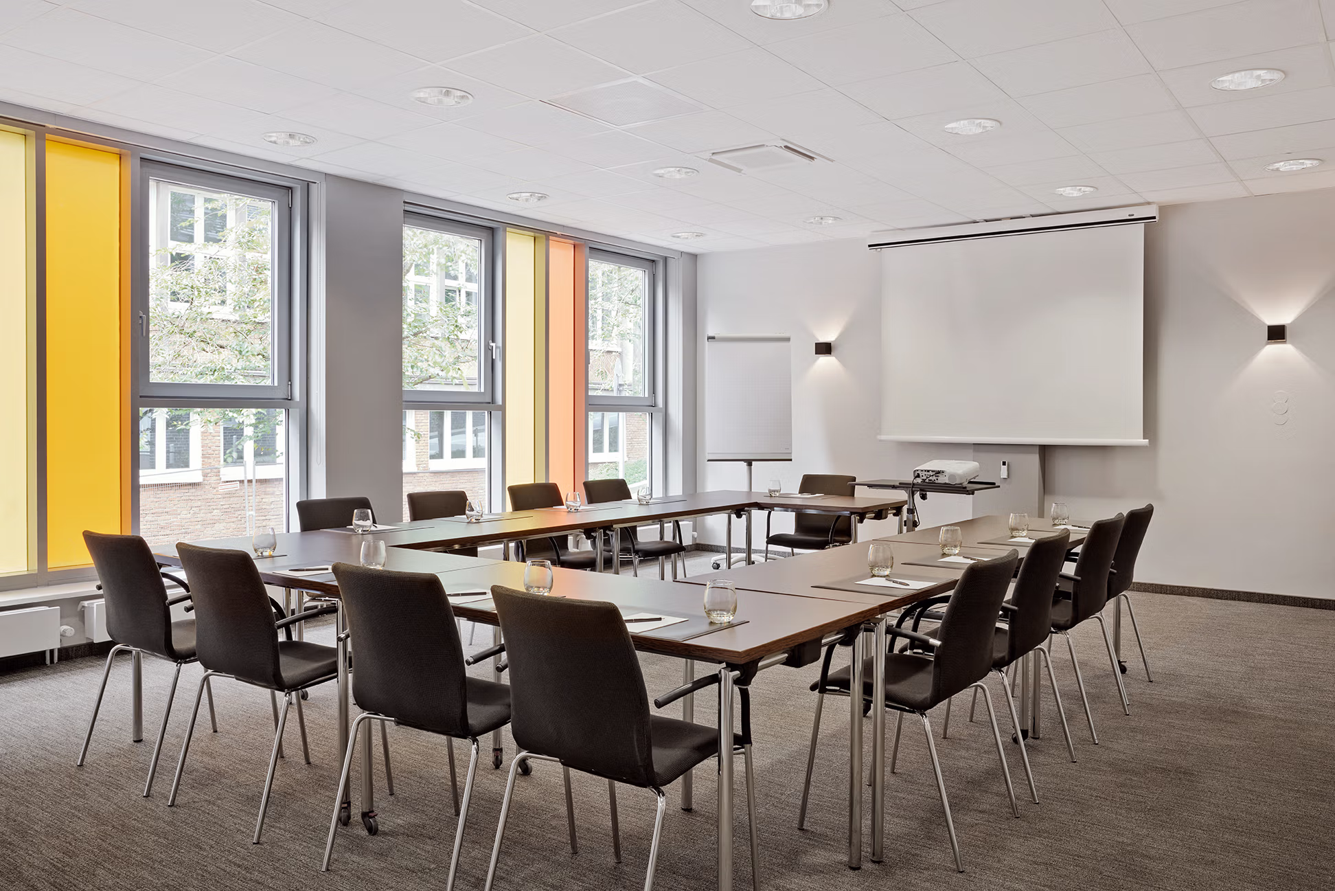 Empty, bright meeting room with U-shaped tables and black chairs; a notepad and a glass of water in each seat. Clean table surfaces in the foreground, a projector in front of a large screen and a flip chart on the wall. Large windows with yellow and orange slats let in daylight, neutral walls and discreet ceiling lighting — a quiet, professional atmosphere, ready for a meeting.