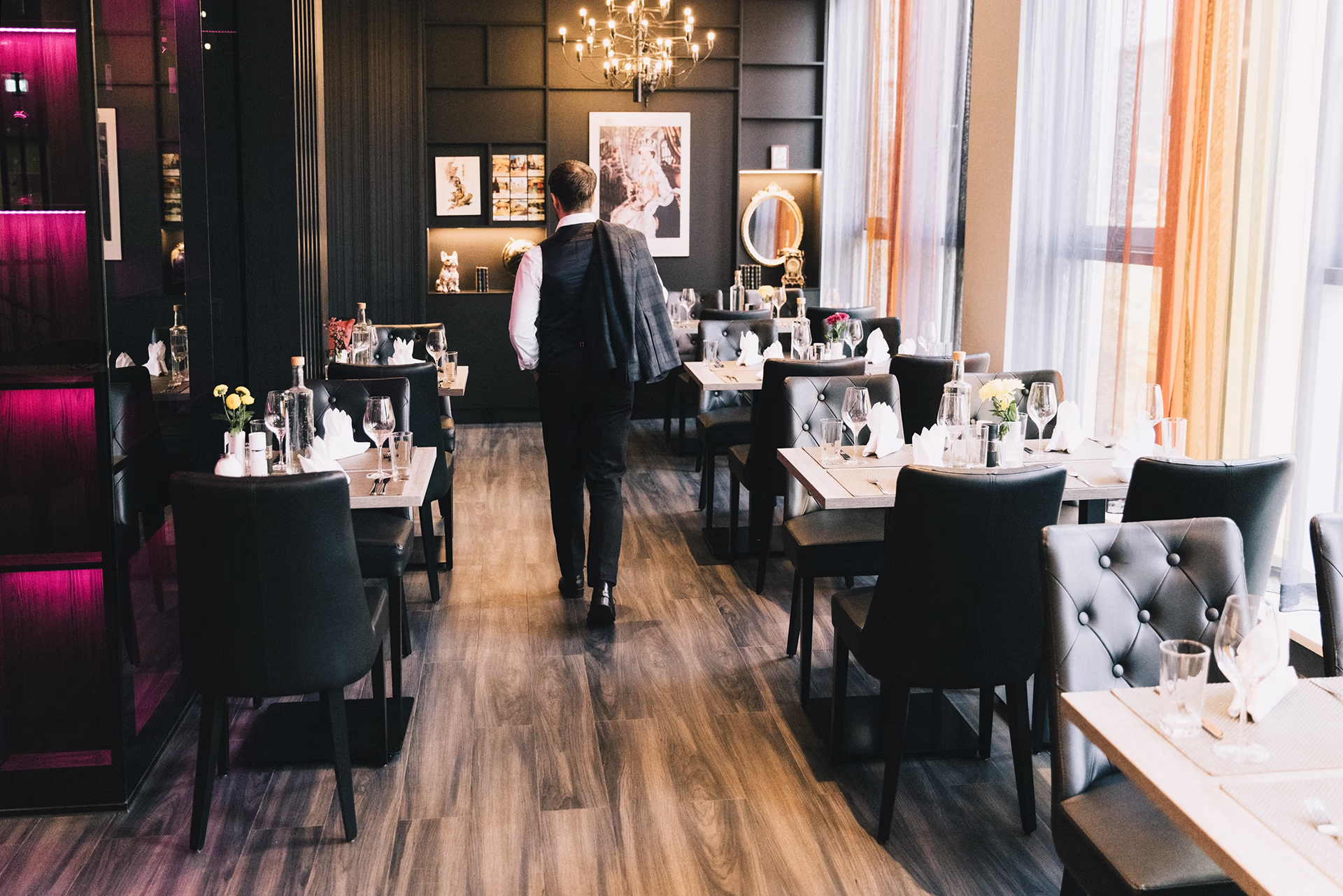 Elegant, warmly lit restaurant with polished wooden floors and a central alley, tables arranged on both sides with black, quilted upholstered chairs. On the set tables, white napkins, wine and water glasses, silverware, small flower vases and water bottles; on the left, a wall of shelves with pink lighting, decorative pictures, a round mirror and a chandelier. A person wearing a shirt and vest wears a jacket over their shoulder and walks to the back — a quiet, muted evening atmosphere, ready for guests.