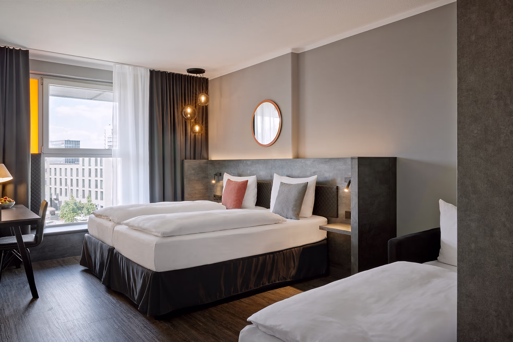 Modern hotel room with two beds — a large double bed in the center and a single bed in the foreground — in calm shades of gray and white; freshly upholstered white sheets, two decorative pillows (one rust-colored, one gray) and dark bed surrounds. An upholstered headboard wall with built-in bedside tables and small reading lamps, above it a round wooden frame mirror and three glassy hanging lamps with warm light. Large windows on the left with white curtains and dark curtains offer views of a city, on the left a desk with lamp and fruit bowl, dark wooden floors and a discreet, cozy hotel room atmosphere.
