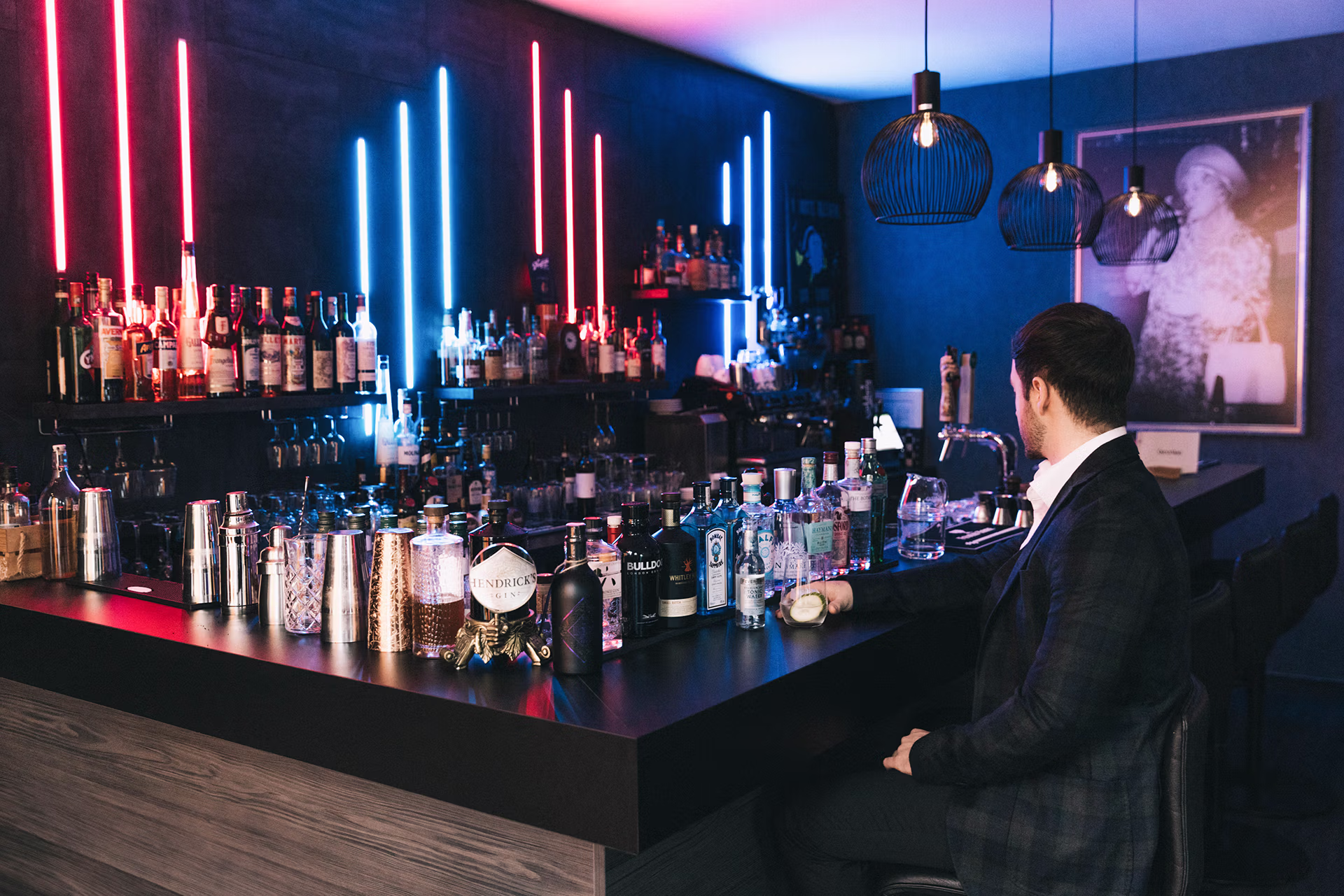 A man in a dark jacket and white shirt sits on a bar stool at a modern bar with vertical neon lights in blue and red, holds a glass with a slice of lime and looks to the side. There are numerous liquor bottles, cocktail shakers and glasses on the black bar counter; hanging wire pendant lights and a framed black and white photo on the wall convey a subdued, cool, nocturnal bar atmosphere.