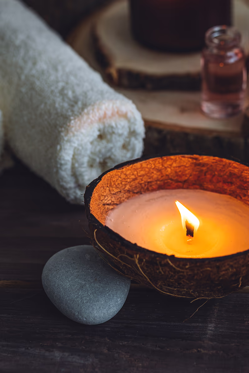 Nahaufnahme einer brennenden Kerze in einer ausgehöhlten Kokosnussschale auf dunklem Holztisch; die Flamme leuchtet warm in geschmolzenem Wachs. Daneben ein glatter grauer Massage-Stein, ein aufgerolltes weißes Handtuch und unscharf im Hintergrund ein kleines Glasfläschchen mit rosa Öl auf runden Holzscheiben – ruhige, entspannende Spa-Atmosphäre.