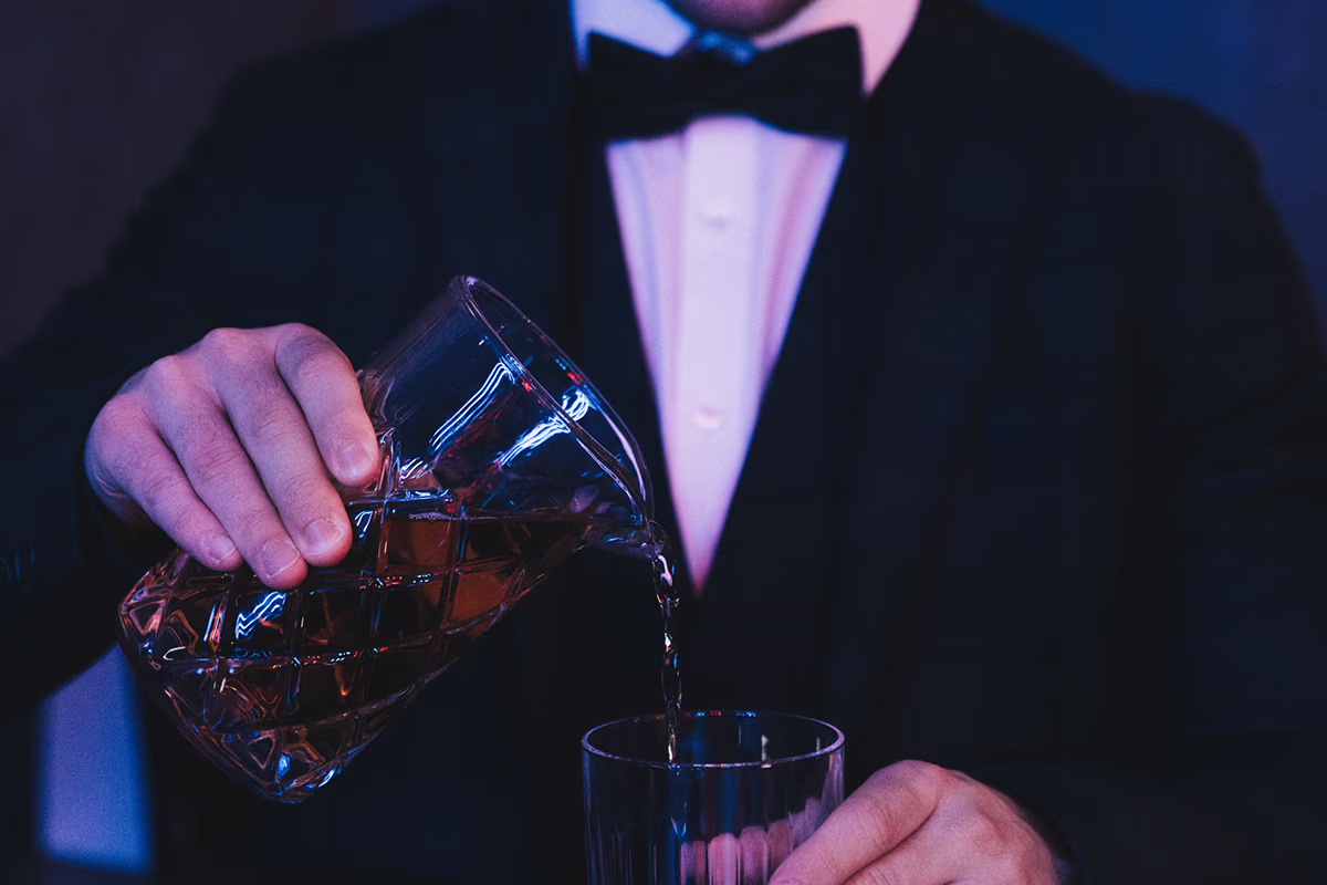 A person in a dark suit with white shirt and bow tie pours amber-colored whiskey from a cut glass pitcher into a tumbler glass; close-up of hands and pouring process, blue-violet muted lighting and elegant, formal mood — concentrated moment when serving a drink.
