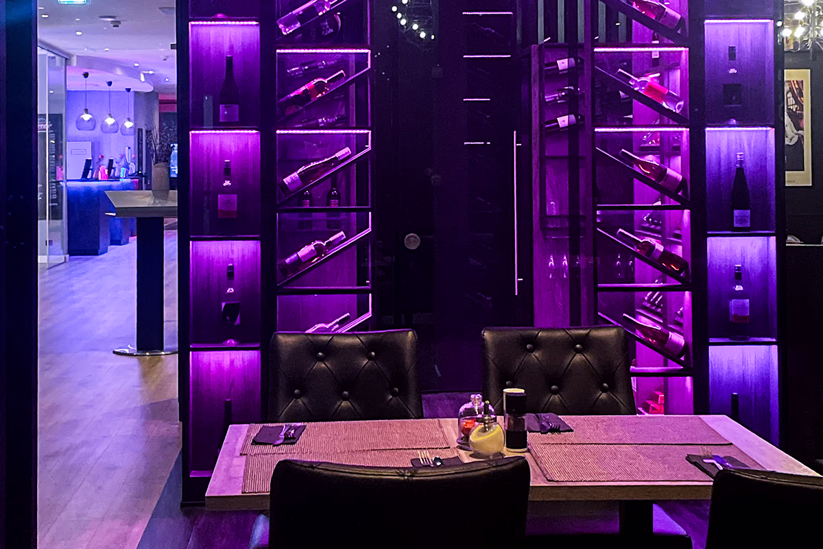 View of the walk-in wine cabinet “The Cube” in the Trinity Restaurant: diagonally set wine bottles behind glass in shelves, dramatically illuminated by purple LED light; in the foreground a table for two with quilted leather chairs, placemats, cutlery and small spice bottles, open left to the hotel foyer with pendant lights and wooden floors — a modern, intimate and stylish atmosphere.