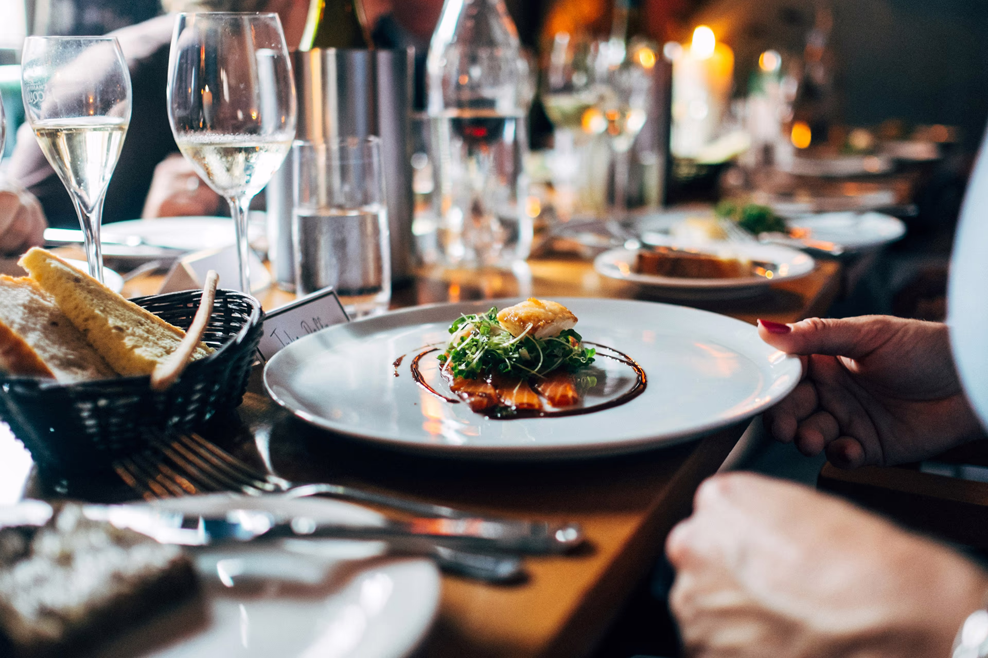 Close-up of a set restaurant table by candlelight: A hand with red nail polish holds a white plate with a small, artfully arranged bite of microgreen on dark dressing; in the foreground cutlery and bread basket with slices of bread, in the background wine glasses with white wine, a water bottle and fuzzy guests, cozy, festive atmosphere.