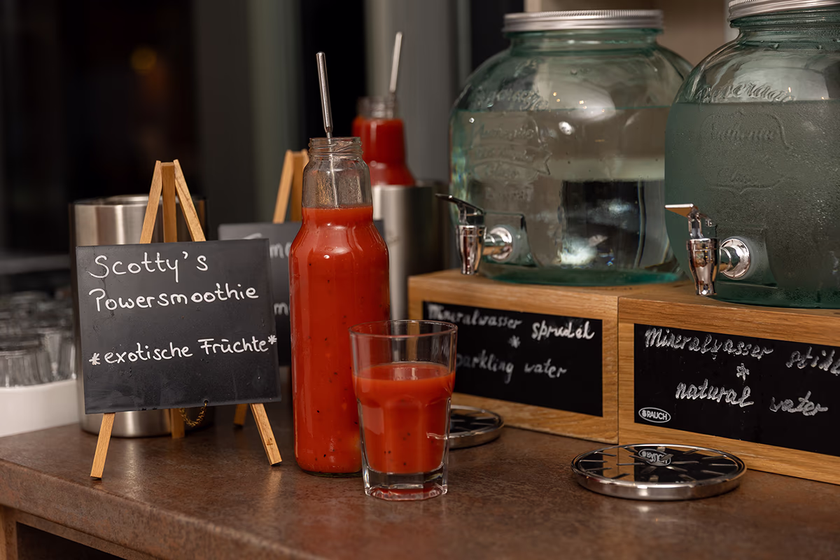 Close-up of a breakfast table in the Scotty Hotel Hamburg: a tall glass bottle with red power smoothie and metal straw is in the middle, in front of it a half-full glass of the smoothie; on the left on a small wooden easel a black chalkboard with the inscription “Scotty's Power Smoothie Exotic Fruits”; in the background a second smoothie bottle, on the right two large bulbous glass water dispensers on wooden frames with chrome-plated taps and inscribed chalkboards. Warm lighting, lightly fogged dispensers and tidy buffet decor create an inviting breakfast atmosphere.