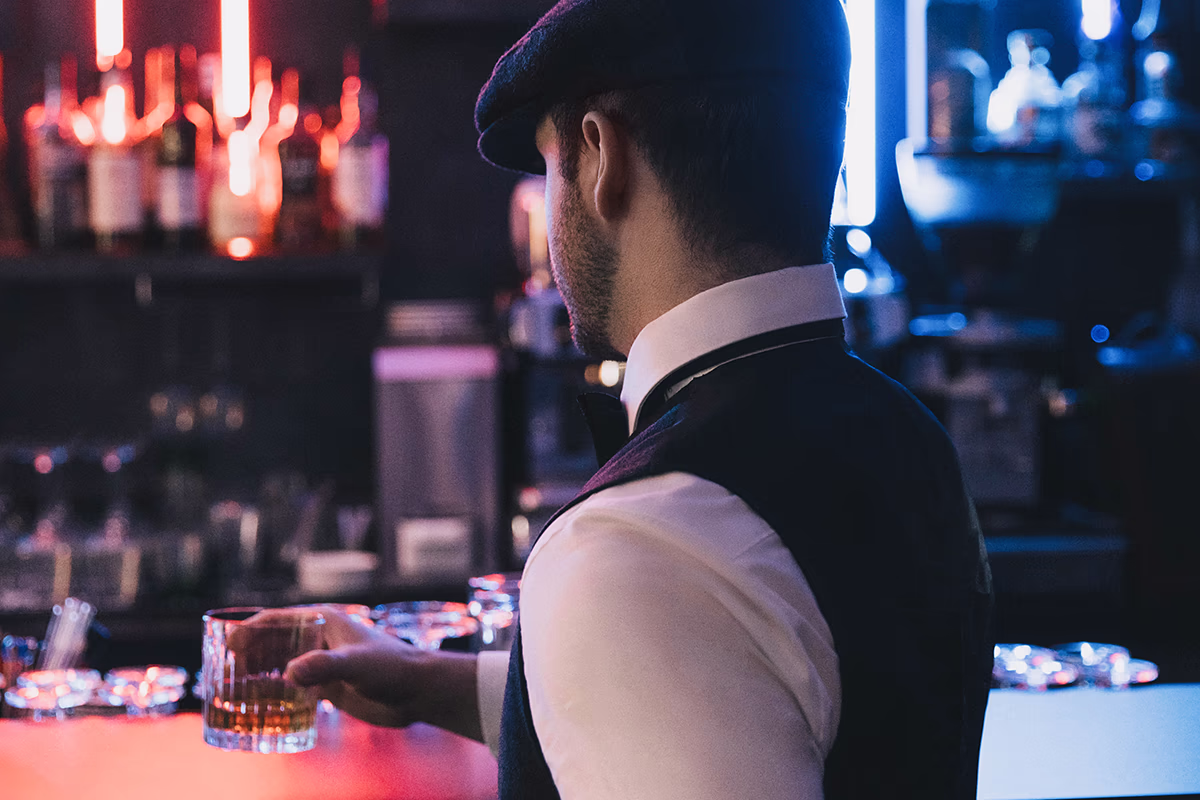 Back view of a person behind the bar in the Queen Mum Bar in the Scotty Hotel Hamburg: the person is wearing a white shirt, a dark vest and a flat cap and holds a faceted glass with whiskey on the bar plate; blurred background with bottles lined up and red/blue neon lighting, subdued, intimate atmosphere.