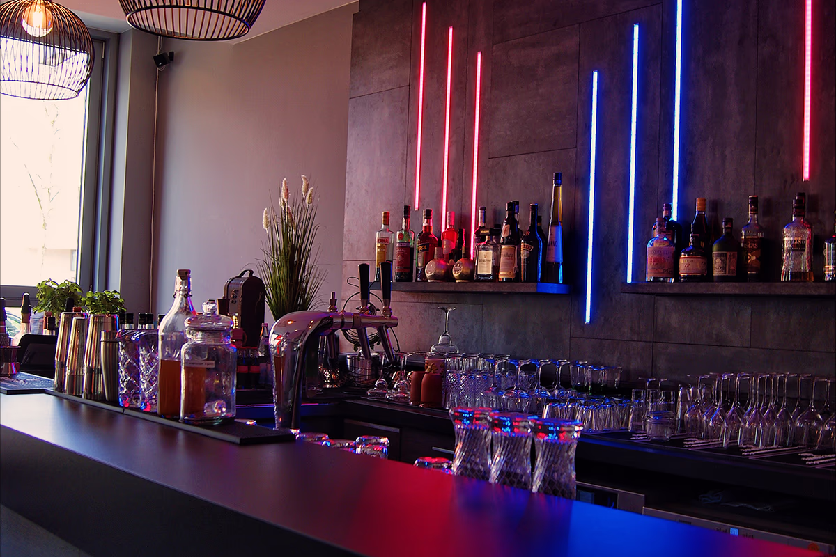 Interior view of the Queen Mum Bar in Scotty Hotel Hamburg: long, dark bar counter with lined up cocktail shakers, glasses and storage glasses in the foreground, taps and wine and spirits bottles on shelves on the back wall; vertical red and blue neon tubes illuminate the scene in contrasting ways and create a modern, atmospheric evening atmosphere, plus hanging basket lamps, a window on the left and a potted plant in the background.