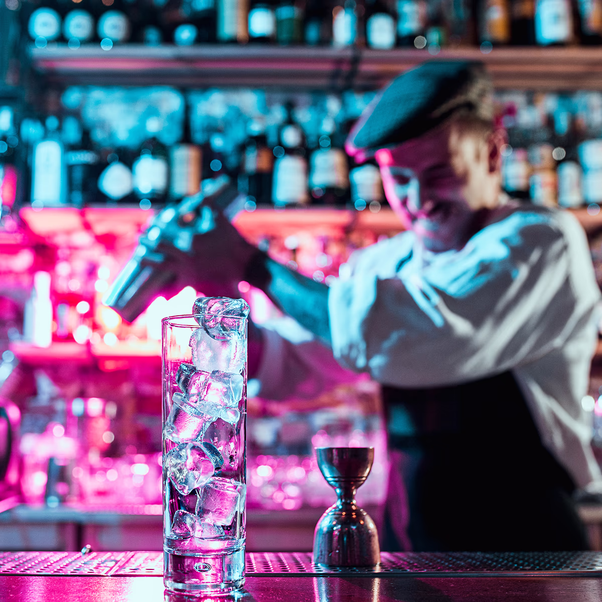 Interior view of the Queen Mum Bar in the Scotty Hotel Hamburg: in the foreground a tall glass full of ice cubes and a metal measuring cup on the counter, in the blurred background a smiling bartender with a flat cap shaking a cocktail shaker; pink and blue neon lights and blurred bottle racks create a lively, lively atmosphere.
