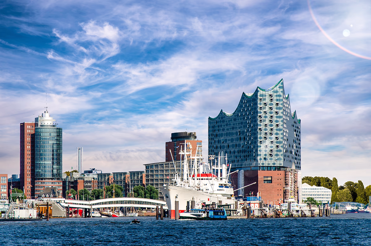 Blick von der Elbe auf die Hamburger Elbphilharmonie: modernes, wellenförmiges Glasgebäude auf rotem Backsteinpodest am Hafen, davor ein weißes Schiff am Kai, blaues Wasser im Vordergrund und heller, teils bewölkter Himmel mit Sonnenspiegelung, lebhafte Hafenatmosphäre.