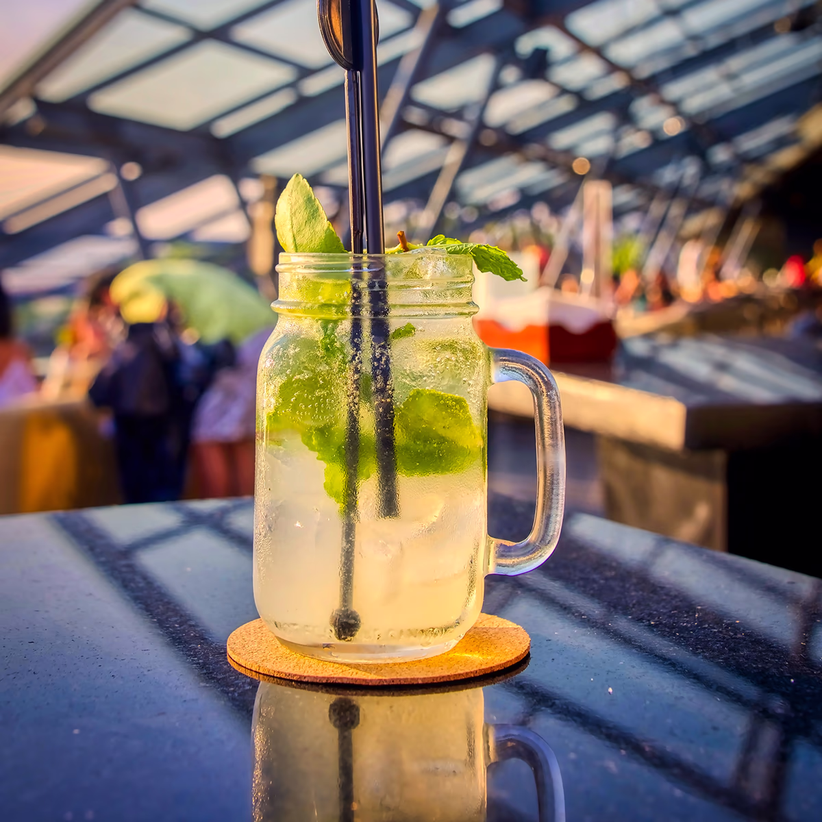 Erfrischendes Minzgetränk mit Eis in einem Einmachglas mit Henkel und zwei schwarzen Strohhalmen auf einem Korkuntersetzer, das Glas perlte vor Kondenswasser und spiegelt sich auf dem dunklen Tisch; im unscharfen Hintergrund unter einer gläsernen Dachkonstruktion sitzen Menschen bei warmem Abendlicht und unterhalten sich, entspannte, sonnige Bar-Atmosphäre.