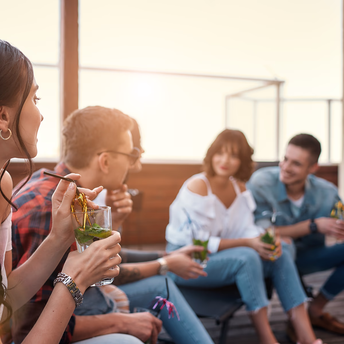 Vier junge Erwachsene sitzen auf einer sonnigen Terrasse in lockerer Runde; eine Frau im Vordergrund hält ein Glas mit Minzgetränk und rührt mit einem Strohhalm, die anderen Personen im Hintergrund unterhalten sich lächelnd; warme Abendsonne, entspannte, gesellige Stimmung.