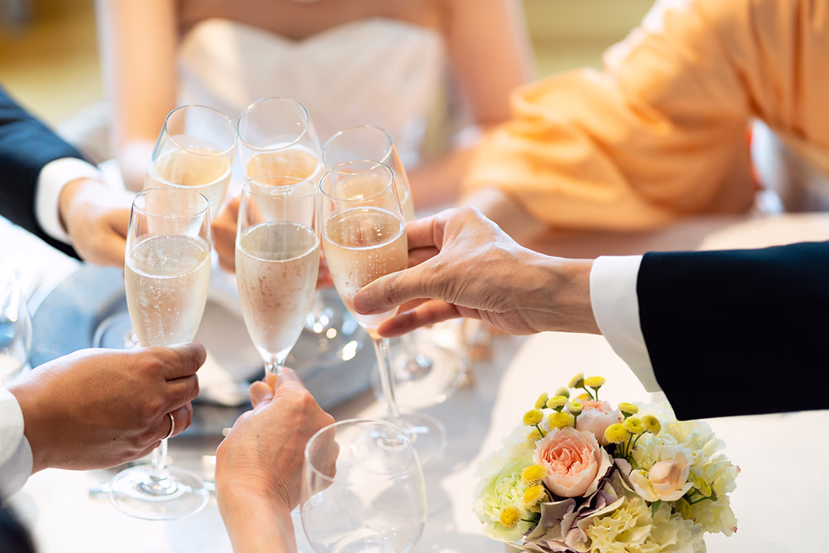 Nahaufnahme mehrerer Hände, die Sektgläser zum Anstoßen heben; im Hintergrund unscharf eine Person im weißen Brautkleid, auf dem Tisch ein zartes Blumenarrangement mit Rosen und gelben Blüten; elegante Kleidung, prickelnder Sekt und warme Lichtstimmung vermitteln festliche, fröhliche Atmosphäre.