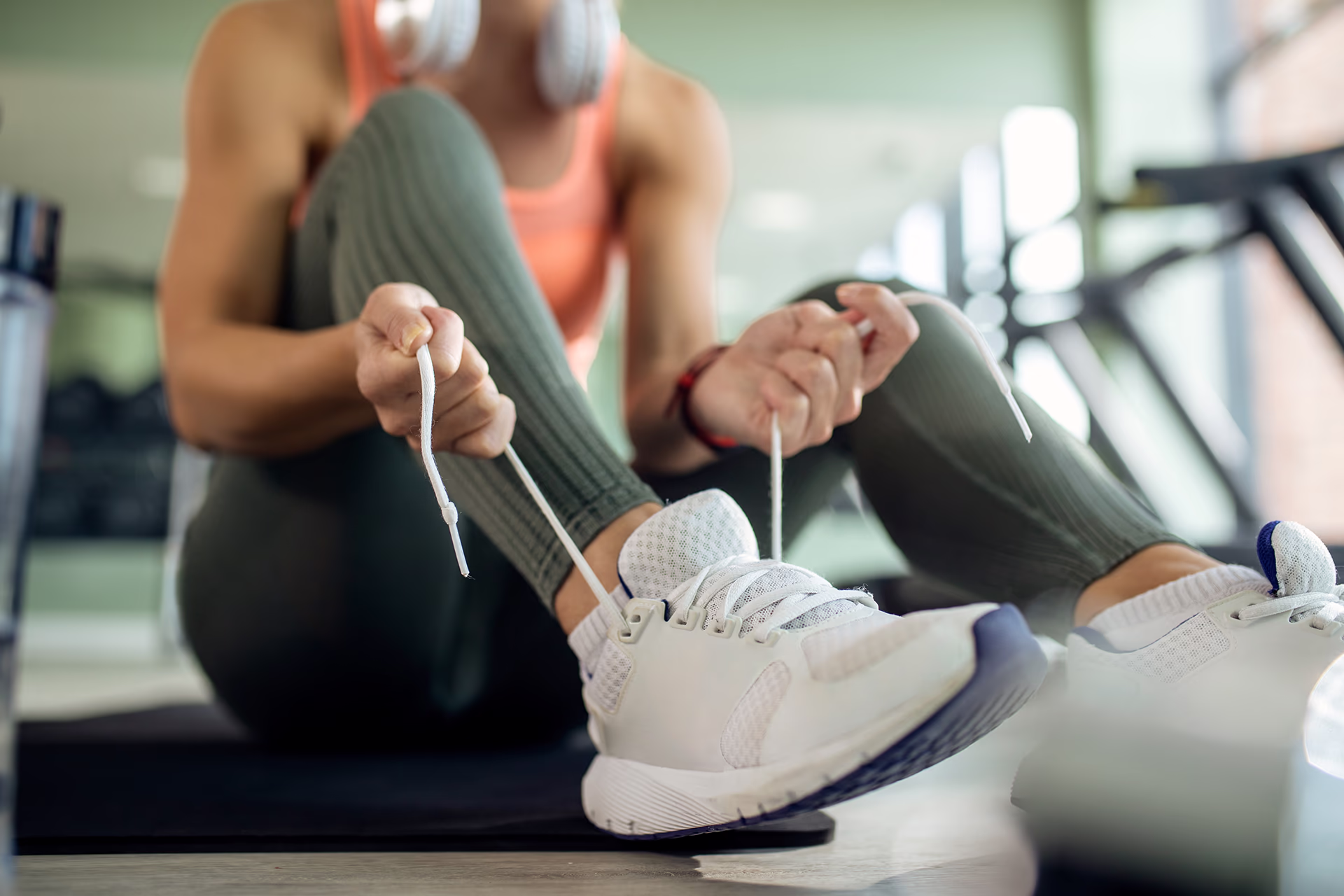 Close up of a seated person lacing white sports shoes: both hands hold white laces over a white running shoe on light ground, the person is wearing green ribbed leggings, a coral-colored tank top and white headphones around their neck and sitting on a black training mat; fitness equipment and a water bottle can be seen in the blurred background. Mood: focused and prepared before training
