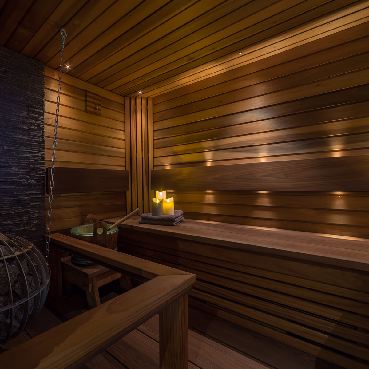 Interior view of an empty light wood sauna with horizontal wooden slats on the wall and ceiling; on the left, a dark slate wall and a sauna stove with stones in a metal basket and a metal chain from the ceiling, in front of it a wooden bucket with ladle on a low bench; on the right, a wide upper bench, towels folded on it and three flickering candles. Warm, diffuse recessed lights create a calm, relaxing spa atmosphere.