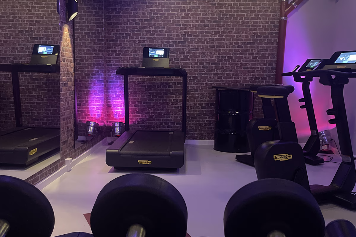 Empty gym with focus on a black treadmill in front of a brick wall, a large mirror on the left, two stationary bicycles/ergometer with screens, purple-pink mood lighting on the wall, in the foreground two large round weights (dumbbell balls or medicine balls) and a black shape-like piece of furniture; clean bright floor, quiet and subdued atmosphere.