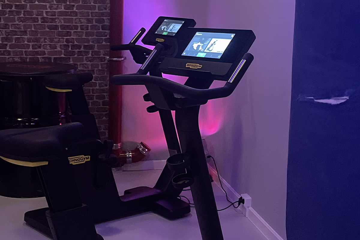 Two black stationary bicycles/ergometers with touch screens in a corner of a gym; purple-pink mood lighting casts light on the wall, red pipeline and brick wall in the background, a shiny black keg as a shelf on the left, visible power cables and power outlet on the floor, quiet and modern training atmosphere.
