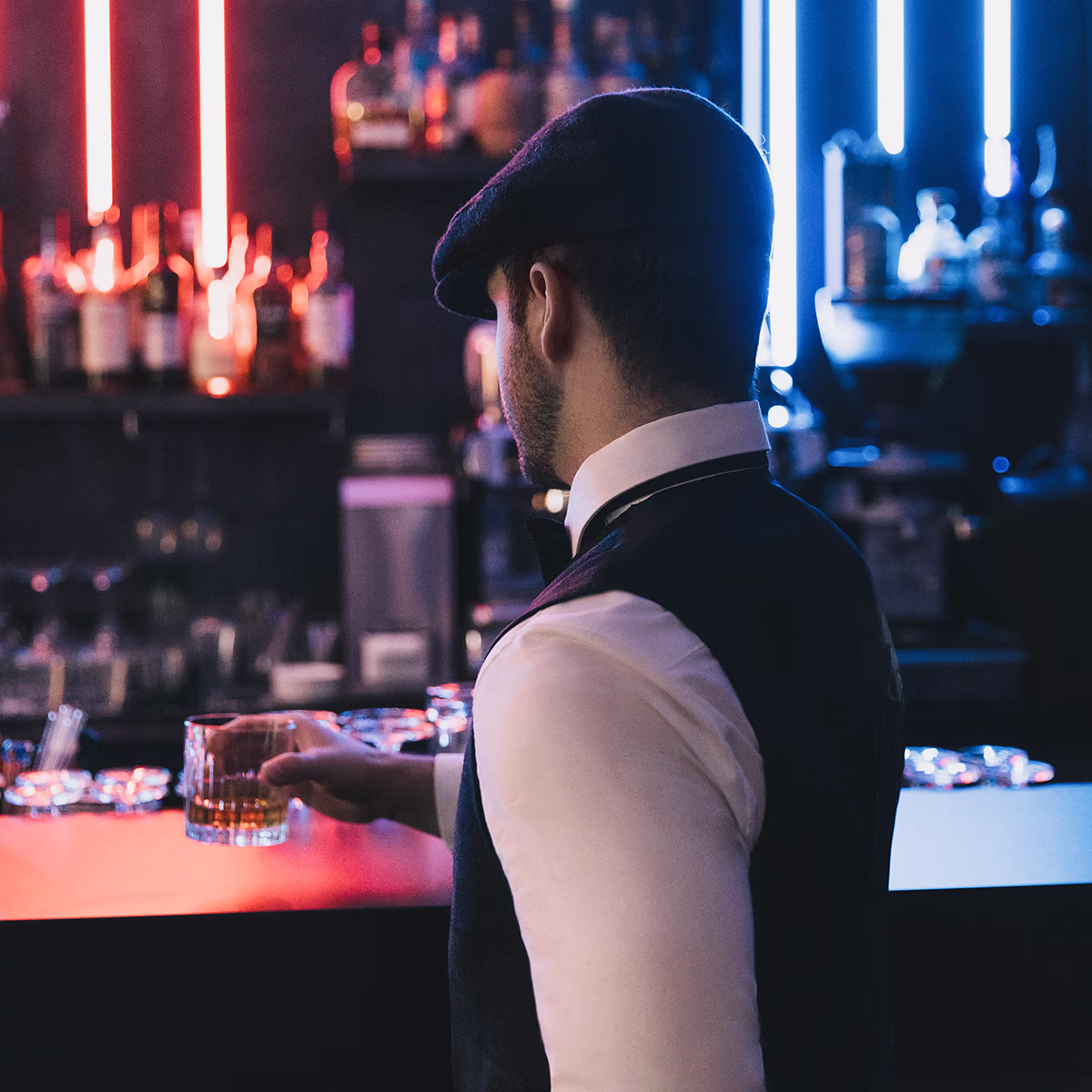 A person wearing a flat cap, white shirt and dark vest sits at a bar, taken from behind and holding a glass of whiskey on the counter; blurred bottles and red and blue neon lights in the background, cozy, dark, nostalgic atmosphere.