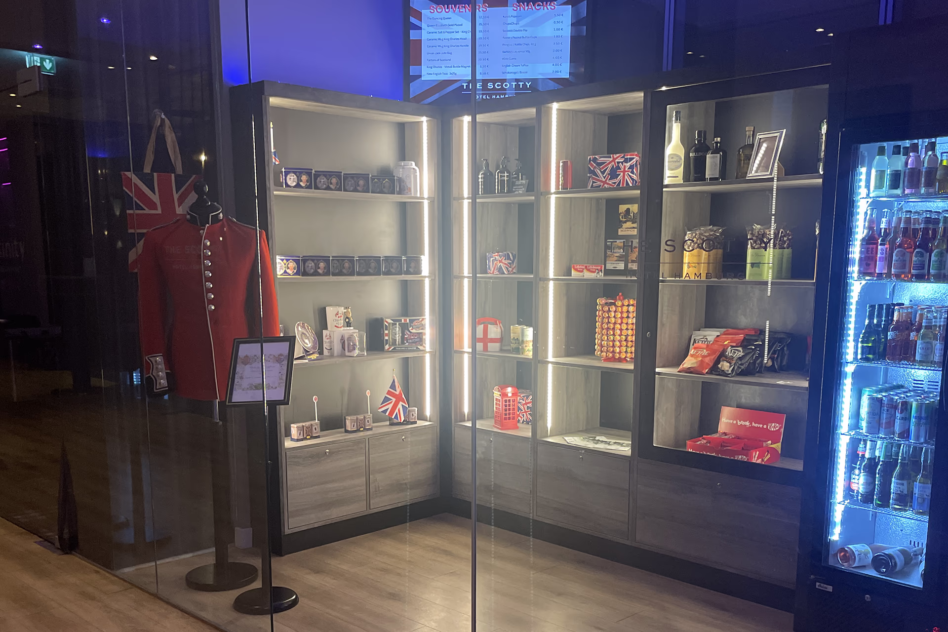View of the small shop of the Scotty Hotel Hamburg through a glass front: on the left a shop window mannequin with a red garden jacket and a Union Jack pocket, wooden cupboards illuminated in the middle and right with souvenirs (cups, cans, mini telephone box, Union Jack items), snack packs and spirits, on the right a refrigerated beverage machine with bluish LED light. The room is tidy and modern, British-themed; the combination of warm shelf lights and cool refrigerator lighting creates an inviting, contemporary ambiance.