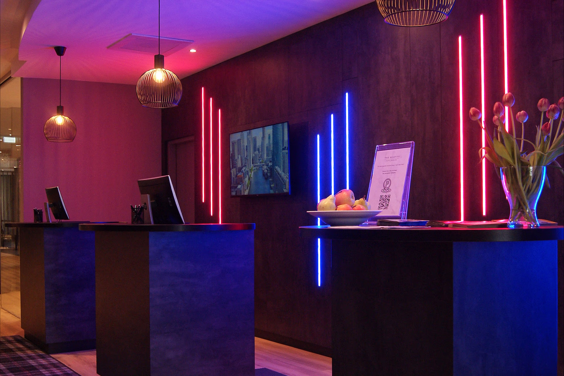 Modern hotel lobby with three black reception desks, two computer screens on the left and a smaller bar on the right. Dark wall in the background accentuated by vertical neon lights in pink and blue, above it two round wireframe hanging lamps, on the wall a screen with a city motif. On the right counter, a bowl of fruit, an information sheet with a QR code and a glass vase with tulips. Subdued, slightly futuristic ambiance with contrasting warm pink and cool blue light.