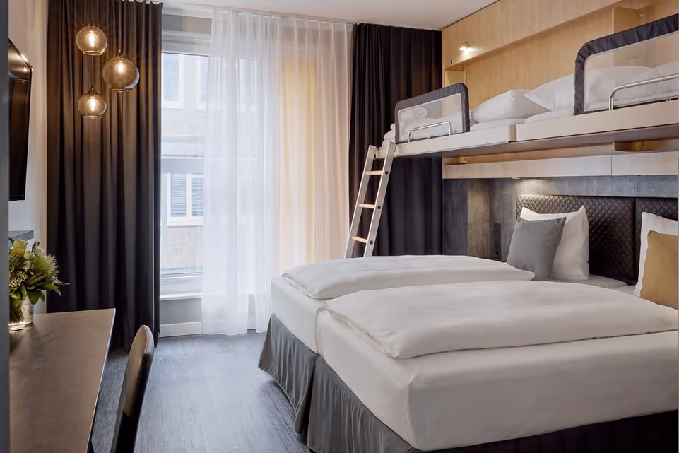 Modernes, warm beleuchtetes Hotelzimmer mit zwei zusammengestellten Einzelbetten im Vordergrund, darüber ein erhöhtes Etagenbett mit Leiter und Sicherheitsgitter; gestepptes dunkles Kopfteil, mehrere Kissen und dezente Lesebeleuchtung; links ein Schreibtisch mit Stuhl, Fernseher an der Wand und ein kleiner Blumenstrauß; großes Fenster mit weißen Gardinen und dunklen Vorhängen sowie drei hängende Kugel-Pendelleuchten. Saubere, ruhige und familienfreundliche Atmosphäre in Holz- und Grautönen.