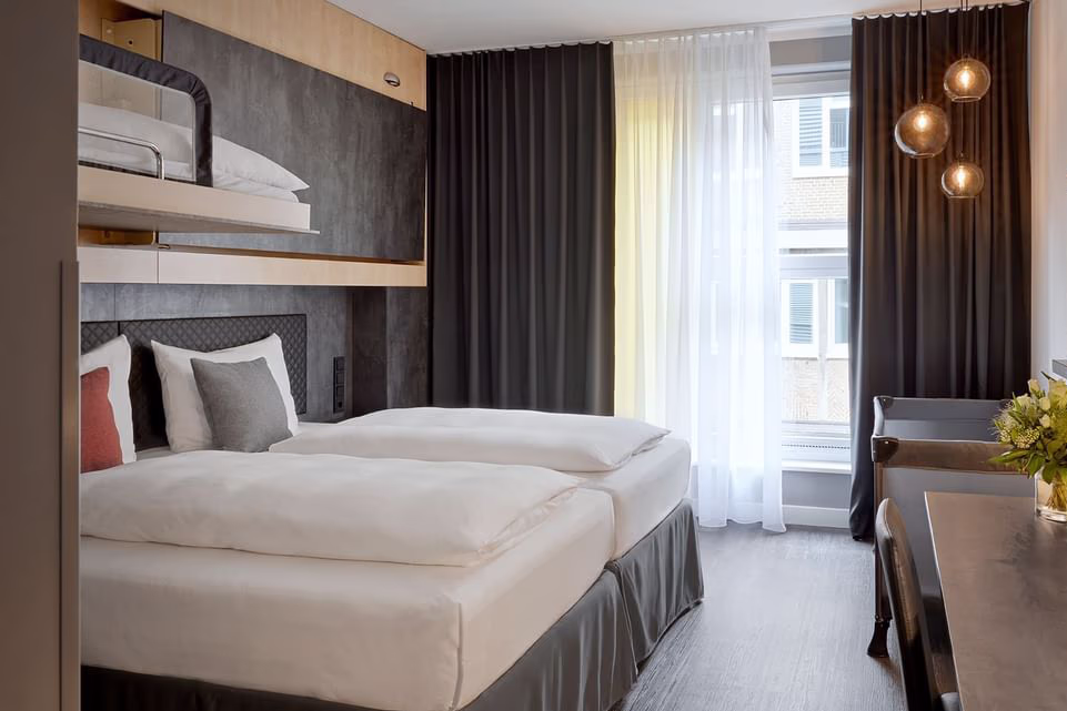 Helles, modernes Hotelzimmer mit zwei zusammengestellten Einzelbetten in weißer Bettwäsche, jeweils weißen Kopfkissen und dekorativen Kissen (links rostrot, rechts grau); darüber ein fest eingebautes oberes Etagenbett mit Geländer; dunkles, gesteppstes Kopfteil vor anthrazitfarbenen Wandpaneelen und Holzleisten; großes Fenster mit weißen Gardinen und dunklen Vorhängen lässt Tageslicht einfallen; rechts drei hängende runde Glaslampen und ein Schreibtisch mit Vase und frischen Blumen. Ruhige, warme und gepflegte