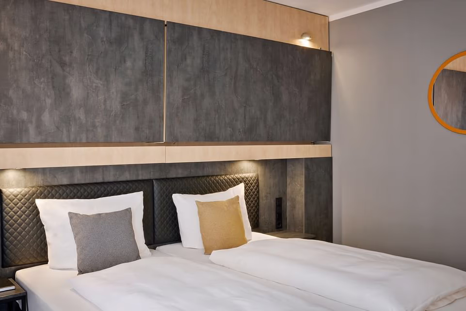 Zweibett-Hotelzimmer mit zwei nebeneinander stehenden Einzelbetten in weißer Bettwäsche, jeweils einem weißen Kopfkissen und einem dekorativen Kissen (links grau, rechts senfgelb); dunkles, gesteppstes Kopfteil vor großen anthrazitfarbenen Wandpaneelen mit Holzleisten, integrierte Leselampen und warmes, indirektes Licht; rechts ein runder Spiegel mit orangefarbener Umrandung und eine eingelassene Nische. Ruhige, moderne und gepflegte Atmosphäre.