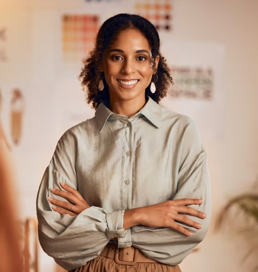Confident woman smiling with arms crossed, standing in a creative workspace with design boards in the background.
