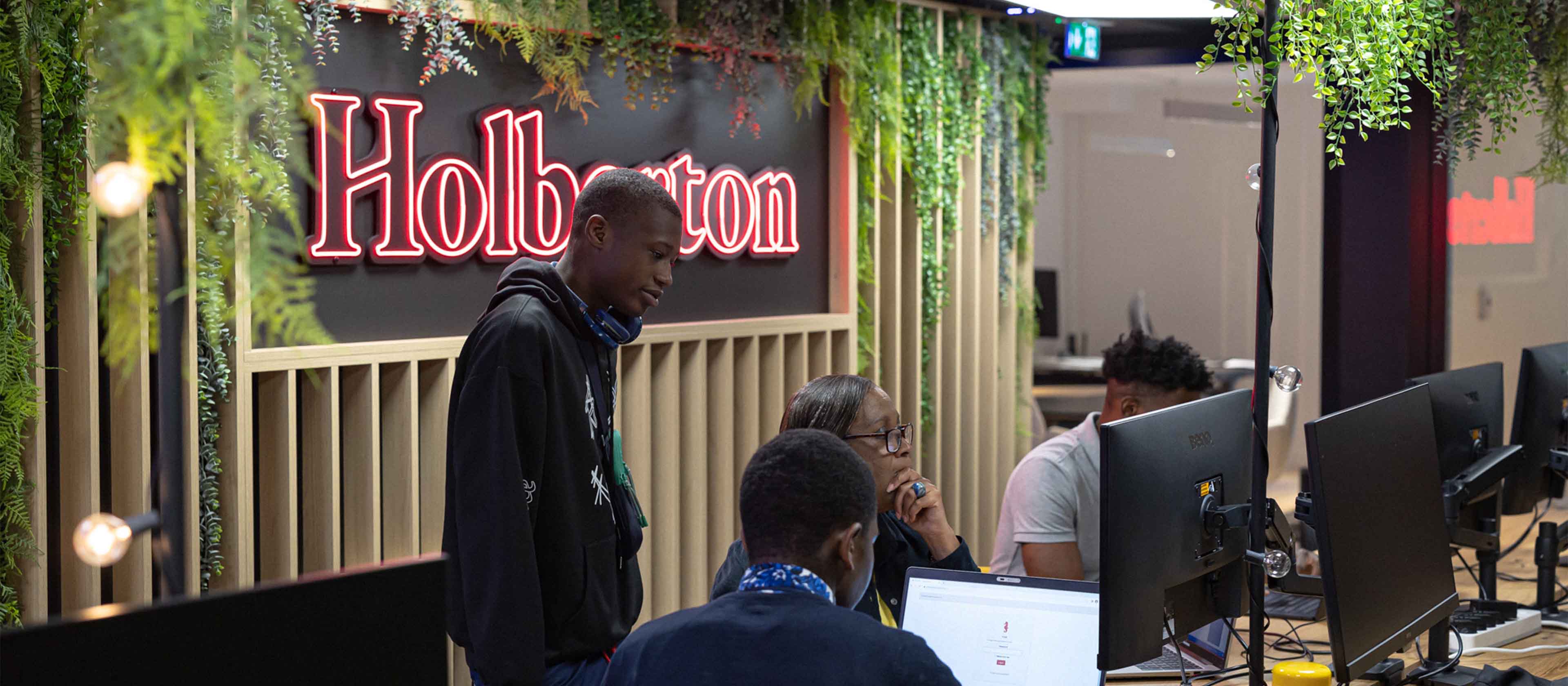 Three people working together in a modern office with computer monitors and a neon Holberton sign on the wall behind them.