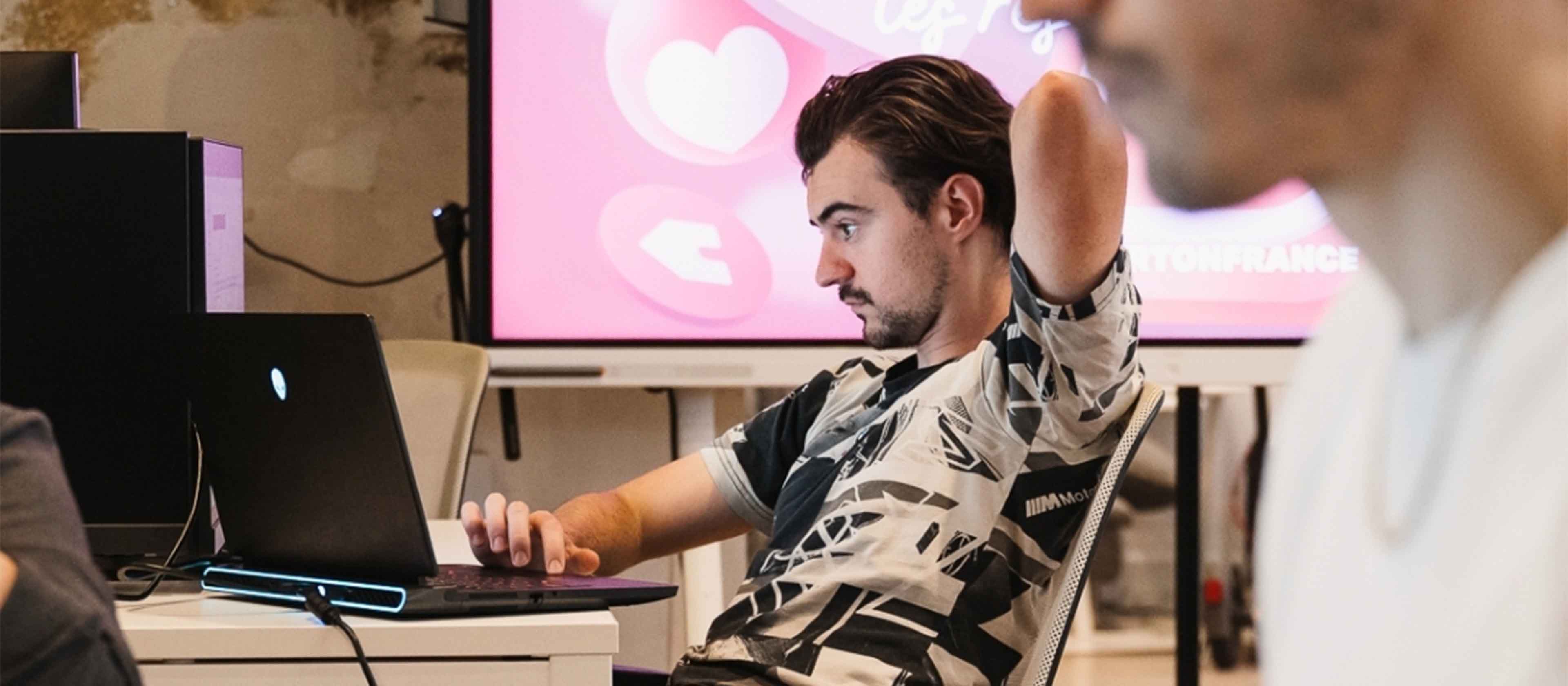Young man sitting at a desk, focused on a laptop with a pink screen displaying heart and arrow icons in the background.