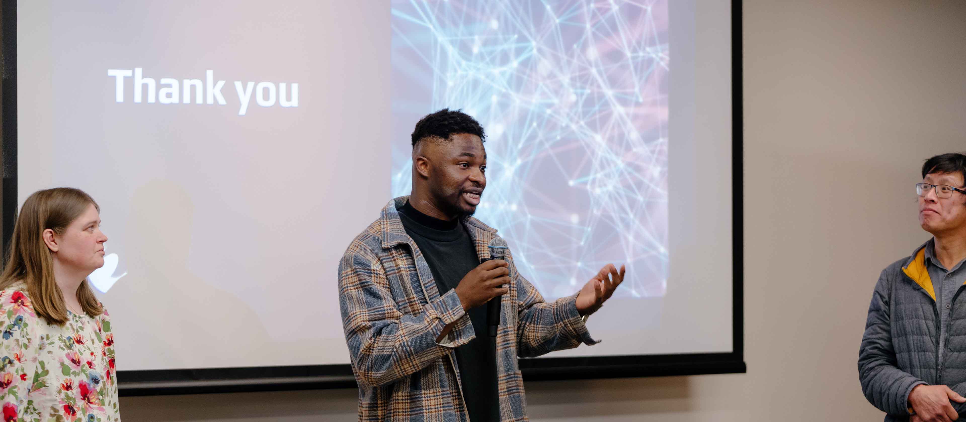 A man holding a microphone speaking in front of a screen displaying the words 'Thank you,' with two people listening attentively on either side.