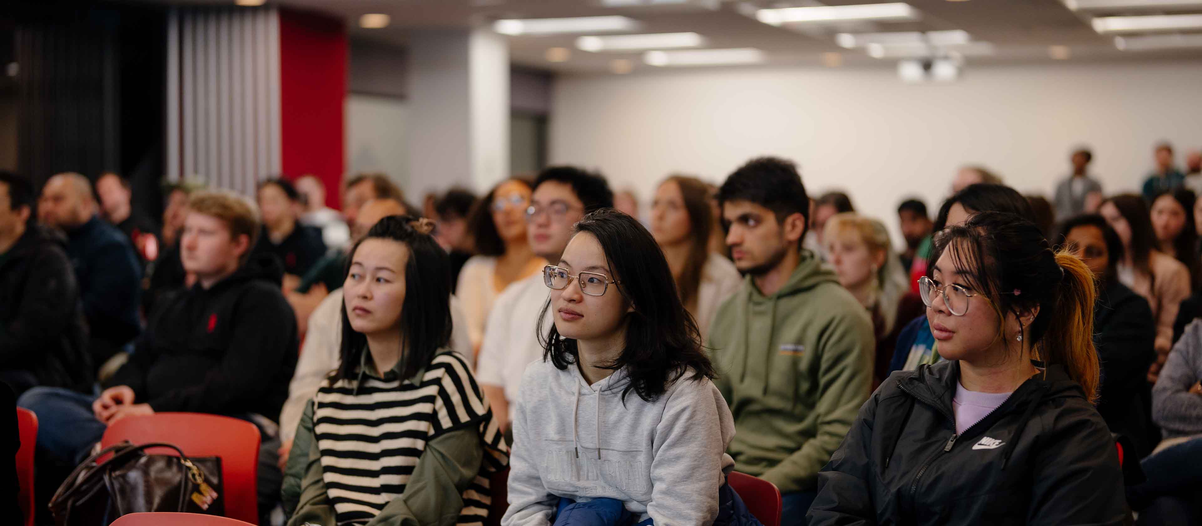 Diverse group of young adults attentively seated indoors, listening to a speaker.