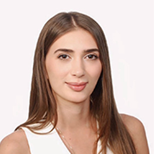 Portrait of a young woman with long brown hair wearing a white top against a plain light background.