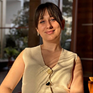 Young woman with brown hair and bangs, wearing a sleeveless cream top, sitting indoors with soft lighting.