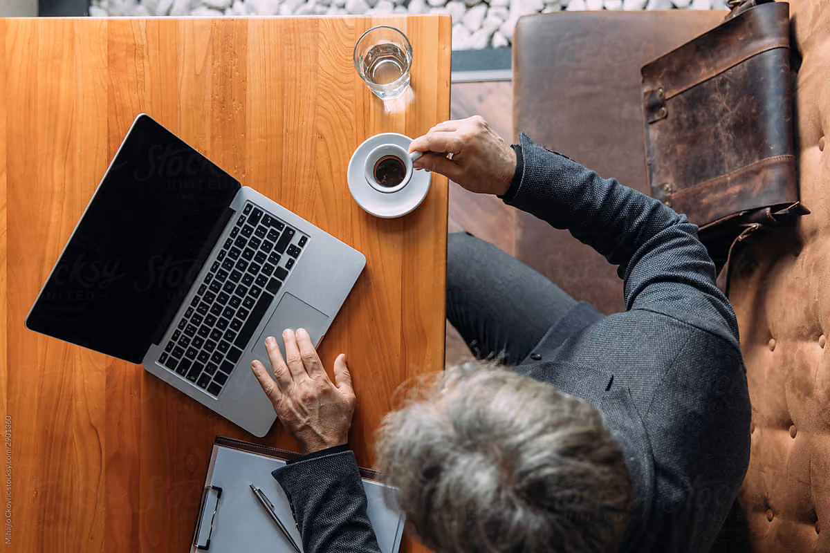 Uomo al tavolo con laptop, tazza di caffè, un bicchiere d'acqua e blocco note con penna.
