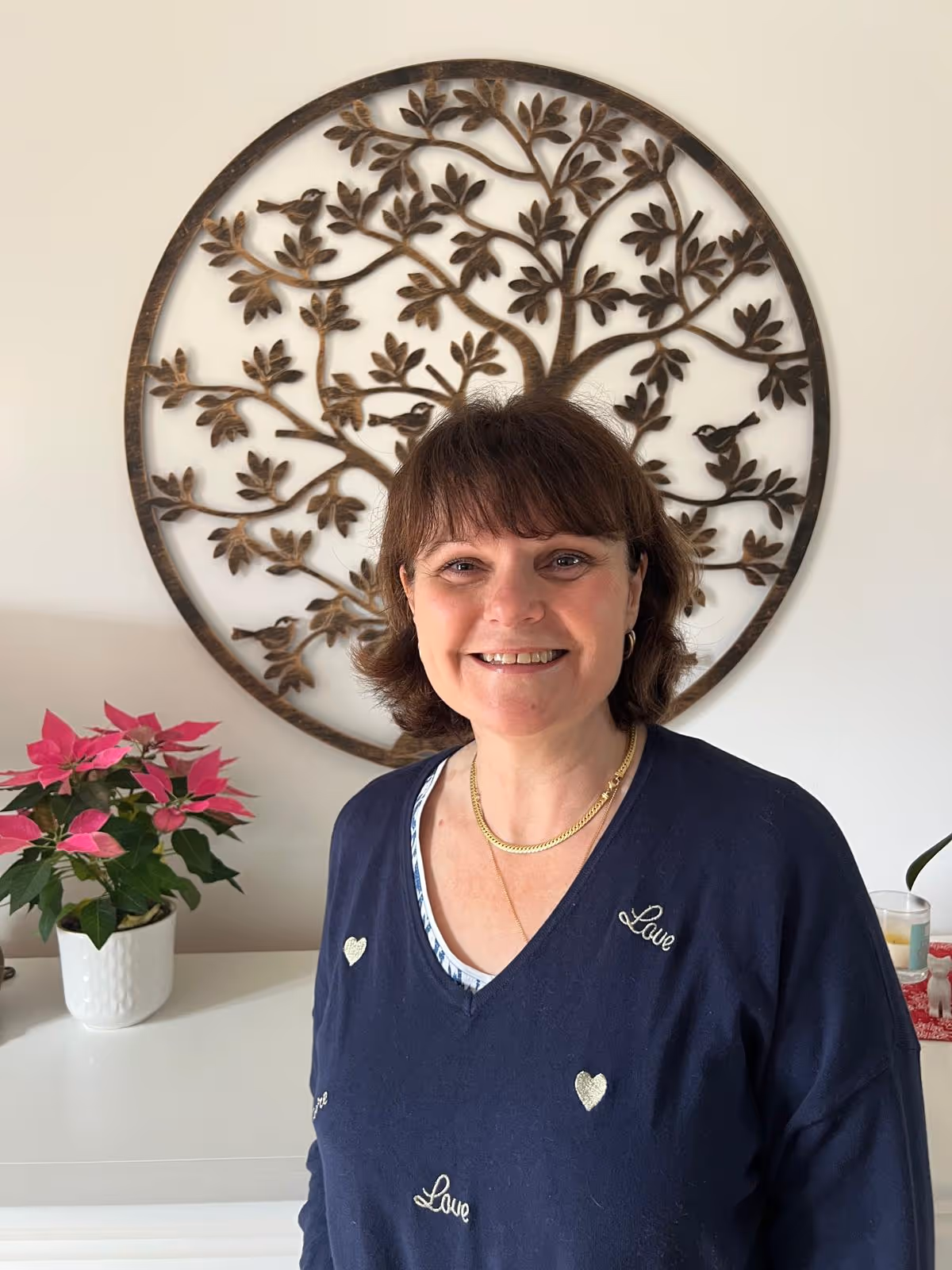 Smiling woman with short brown hair wearing a navy sweater with love and heart designs standing indoors in front of a decorative wooden tree wall art and a poinsettia plant.