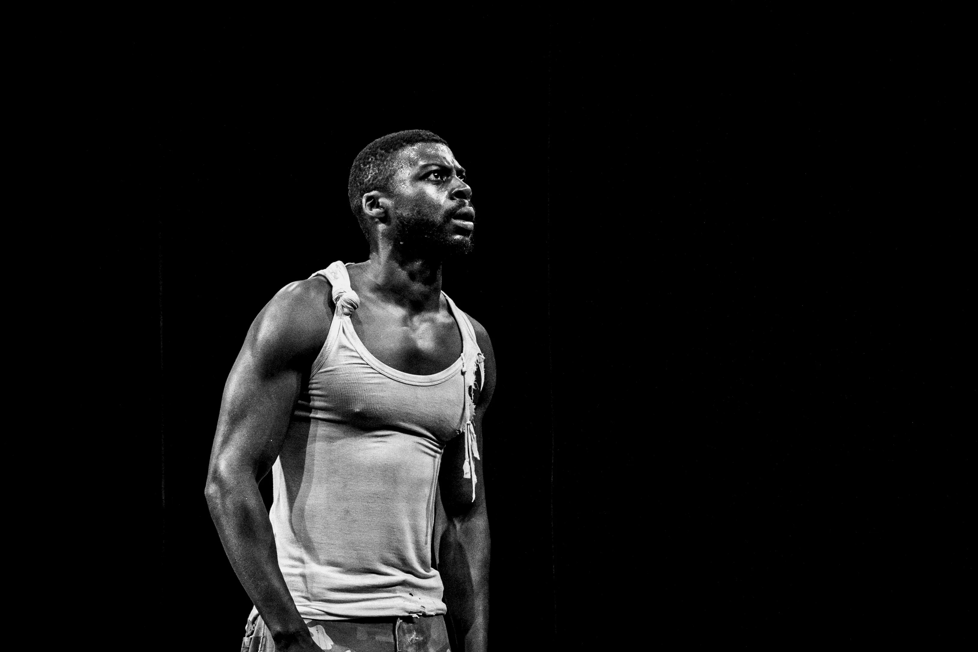 Danseur debout sur scène, regard intense vers le haut, éclairé dans l’obscurité, portant un débardeur blanc et un pantalon déchiré.