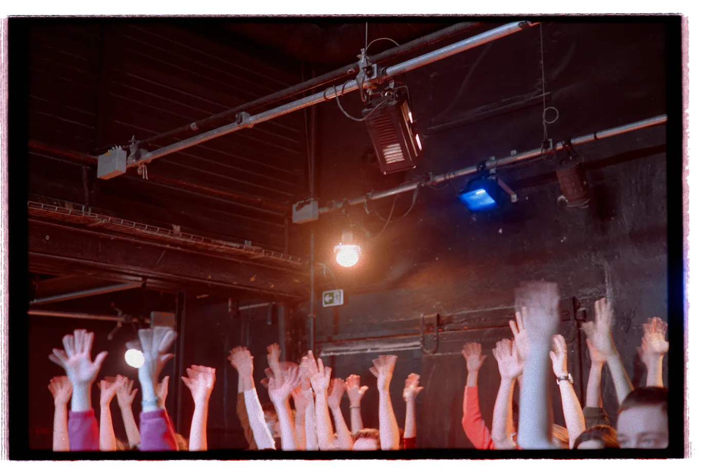 Groupe de personnes levant les bras sous des lumières de scène, dans une salle sombre au plafond industriel.