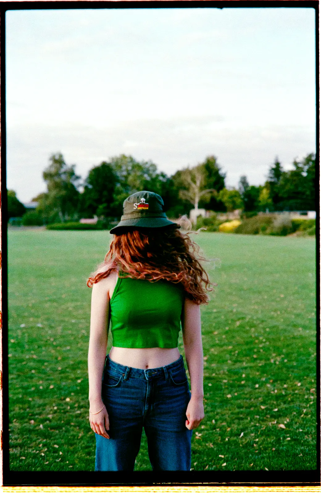 Personne debout sur un champ de verdure, vêtue d’un haut vert et d’un chapeau, les cheveux cachant le visage, dans une lumière naturelle.