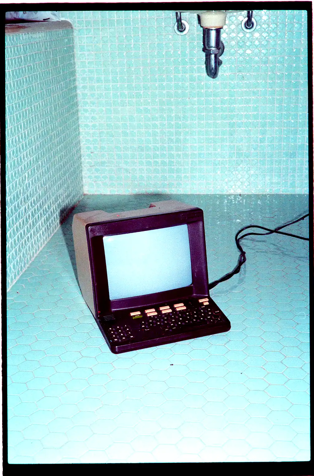 Ancien terminal informatique posé dans un évier carrelé bleu, relié par un câble, dans une ambiance rétro et surréaliste.