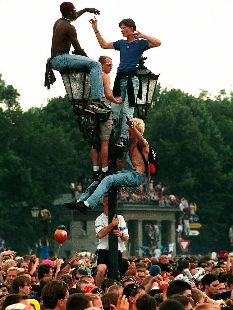 Groupe de jeunes perchés sur un lampadaire au-dessus d’une foule dense en plein air, ambiance de fête ou rassemblement.