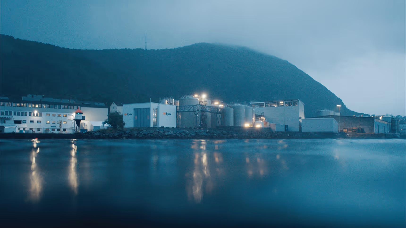 Epax industrial factory complex with large storage tanks and lit buildings by a calm waterfront under a foggy mountain.