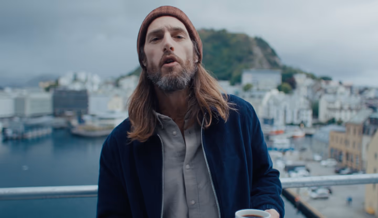 Man with long hair and beard wearing a brown beanie, blue jacket, and gray shirt, holding a cup of coffee with a harbor and buildings blurred in the background.