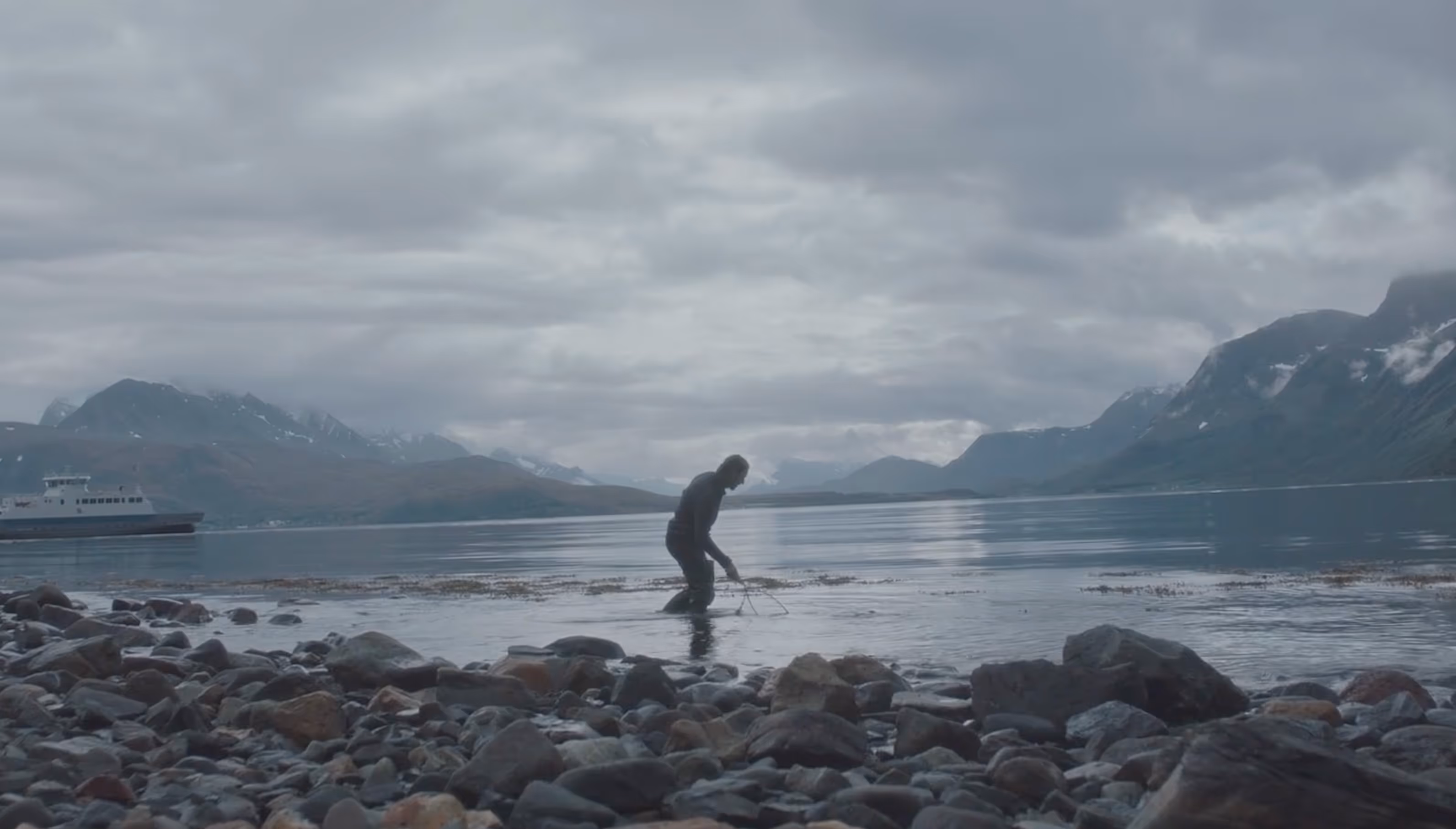 Person standing in shallow water near a rocky shore with mountains and a boat in the background under a cloudy sky.