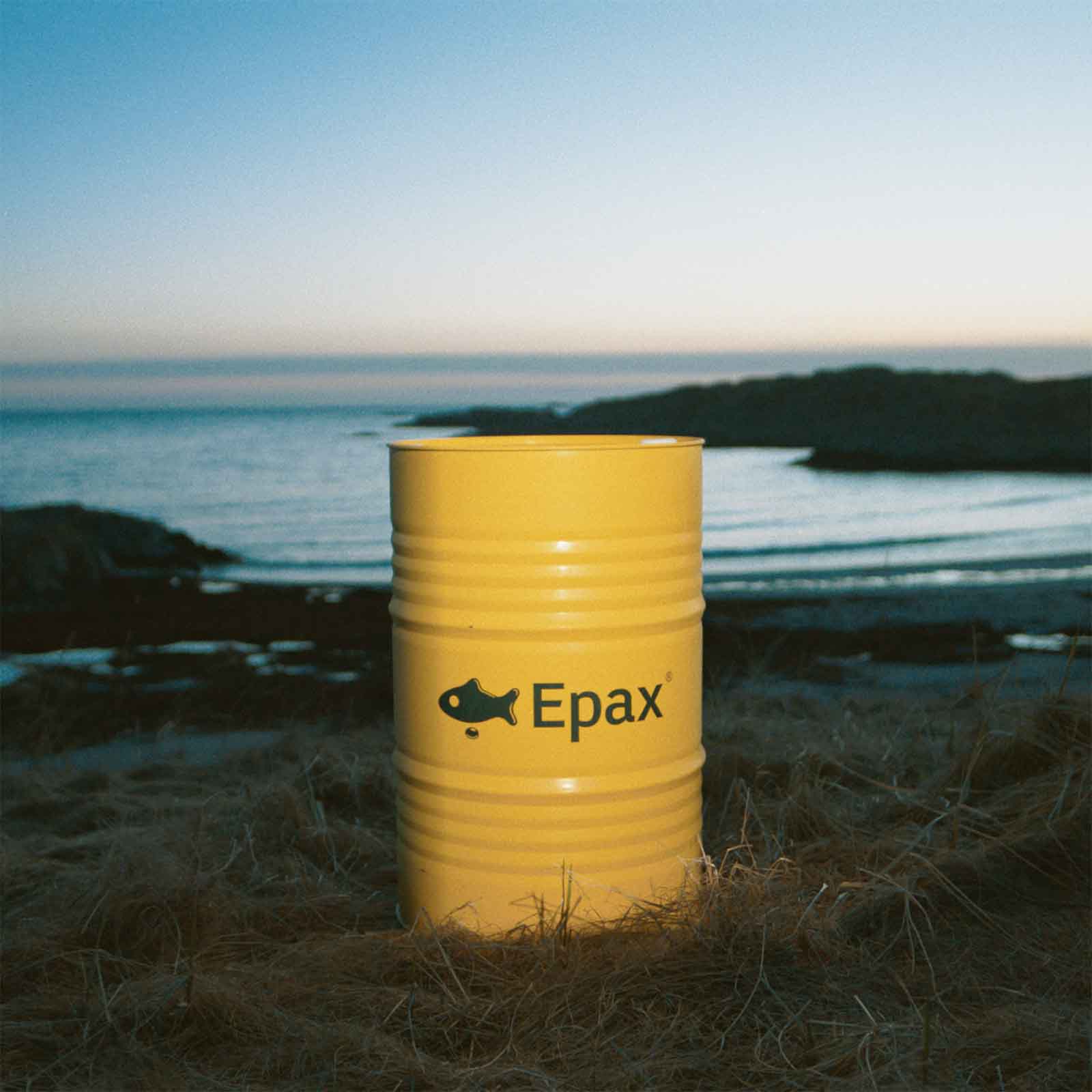 Yellow barrel with Epax logo placed on grassy ground near a rocky shoreline in Karmsund, Norway, at dusk.