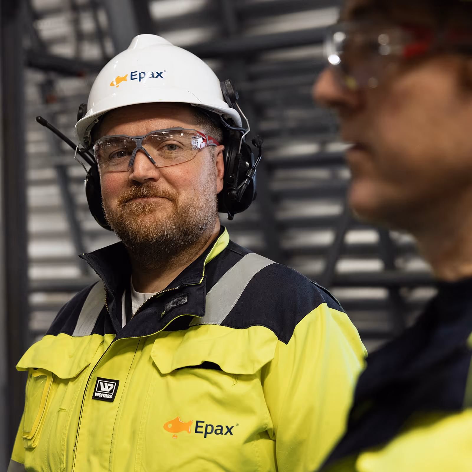 Man wearing a white hard hat and yellow safety jacket with Epax logo, standing in the Epax production facility.