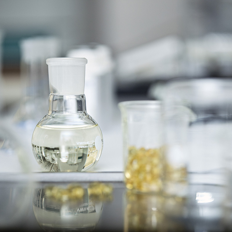 Clear glass flask with fish oil and a white stopper, alongside a beaker containing yellow Omega-3 capsules on a reflective surface in the Epax laboratory.