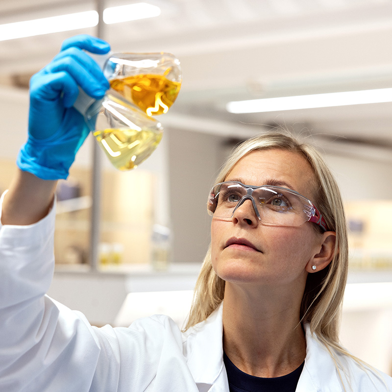 Scientist wearing blue gloves and safety glasses examining two different colored fish oil samples in glass containers.
