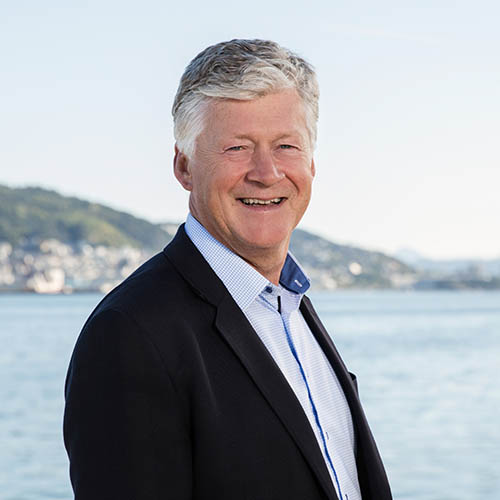 Smiling older man with gray hair wearing a dark blazer and light blue shirt standing by a body of water with hills in the background.
