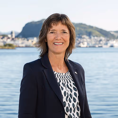 Smiling woman in dark blazer and patterned blouse standing in front of a body of water with blurred mountains and buildings in the background.