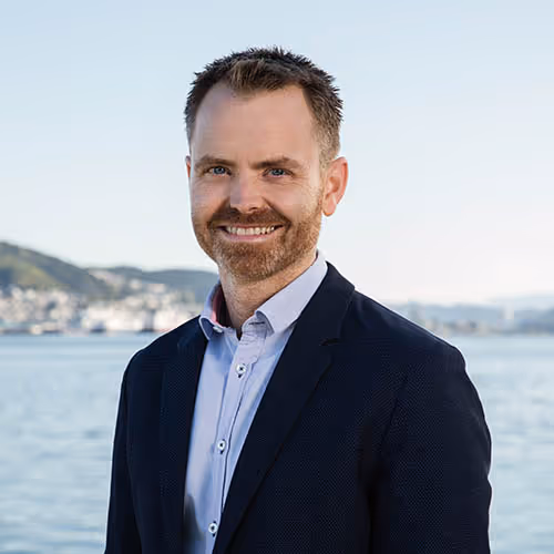 Smiling man with short hair and beard wearing a dark blazer and light shirt standing outdoors near water with hills and buildings in the background.